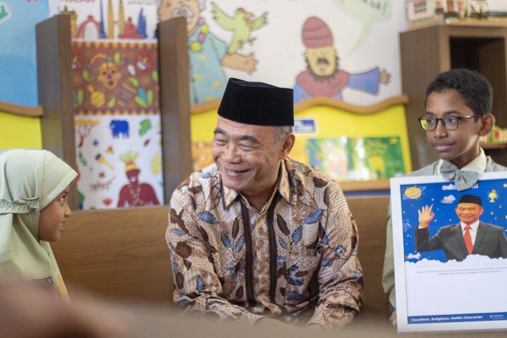 Minister Muhadjir Effendy wearing a bright smile stands in the center. On his right and left, two bright students from Muhammadiyah 4 Elementary School, Anggun and Aby, are also beaming with happiness. They are engaged in an enthusiastic interview at MUPAT Library. Joyful laughter and smiles reflect the cheerfulness and excitement during this moment.
